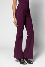 A model wearing high-waisted, wide-leg flared trousers in a deep plum purple color, paired with black lace-up heels, against a light gray studio background.