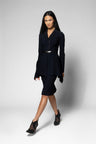 A model wearing a black tailored set consisting of a long-sleeve blazer with a front zipper and a matching knee-length skirt, styled with a thin metallic belt and black high-heeled shoes against a light gray studio background.