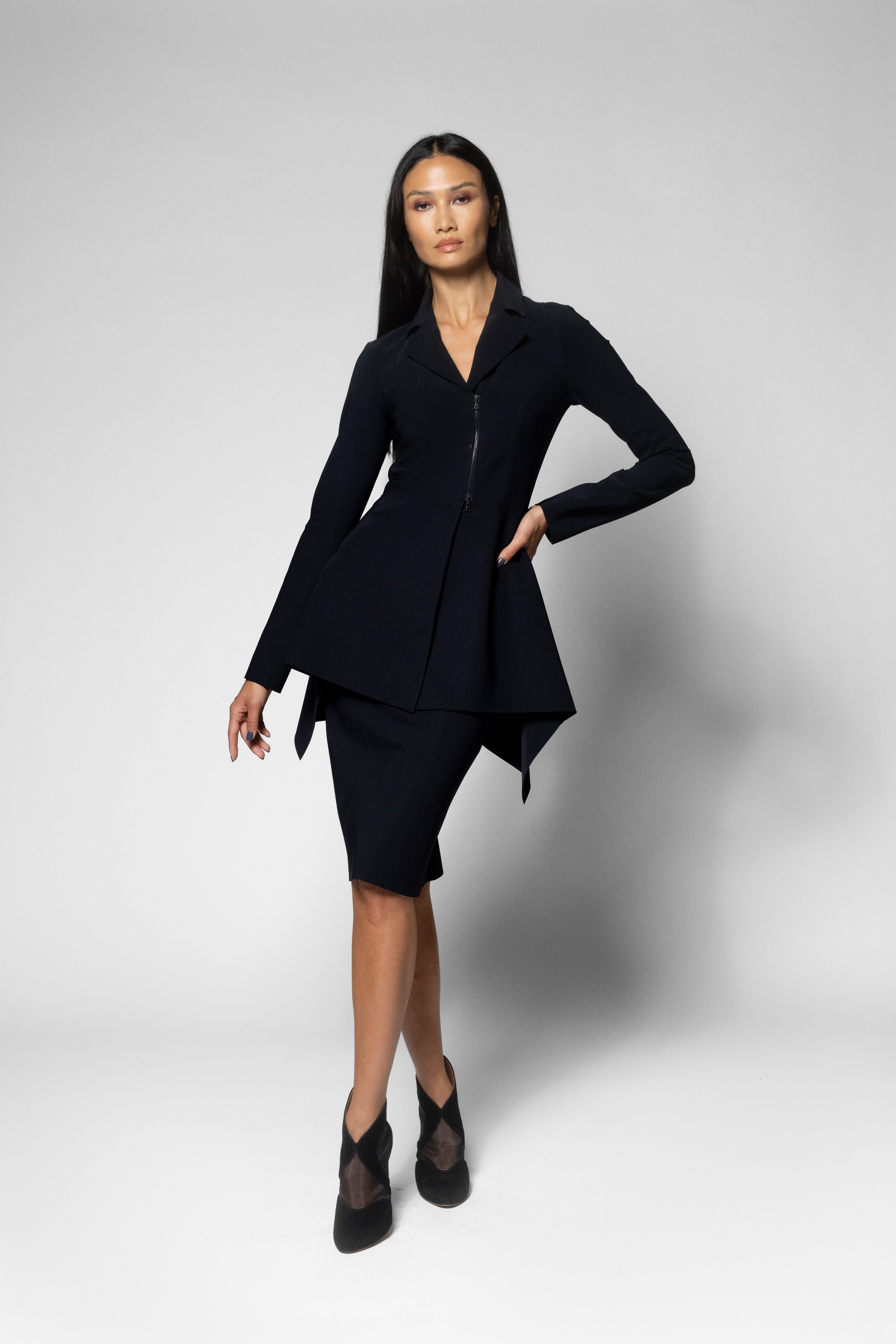 A woman modeling a black tailored two-piece outfit consisting of a long-sleeve peplum-style blazer with a front zipper and matching knee-length skirt, posed against a light gray studio background.