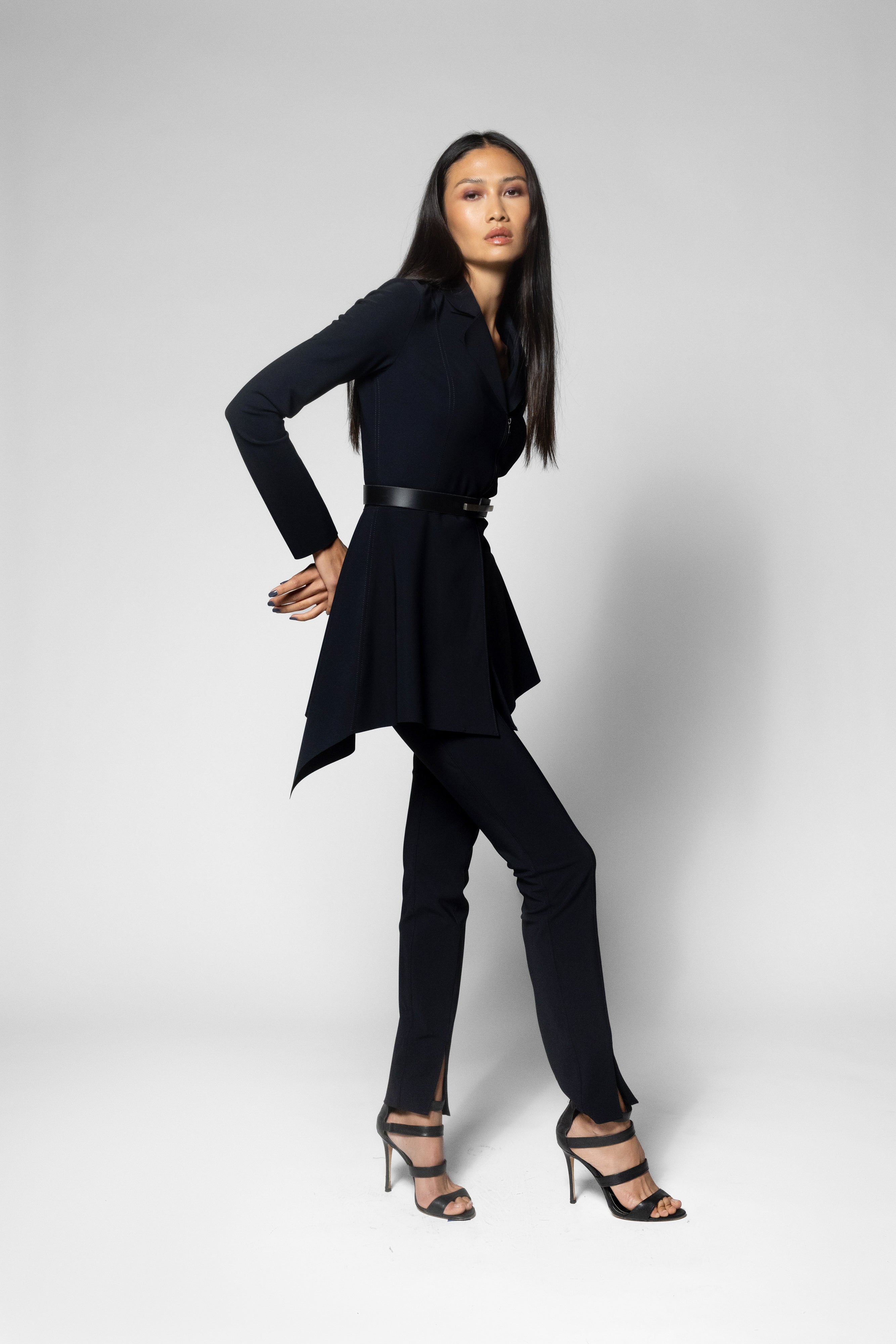 A woman modeling a black long-sleeve wrap-style top with a peplum hem and matching black pants, styled with black strappy high-heeled sandals against a light gray studio background.