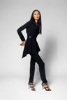 A woman modeling a black long-sleeve wrap-style top with a peplum hem and matching black pants, styled with black strappy high-heeled sandals against a light gray studio background.