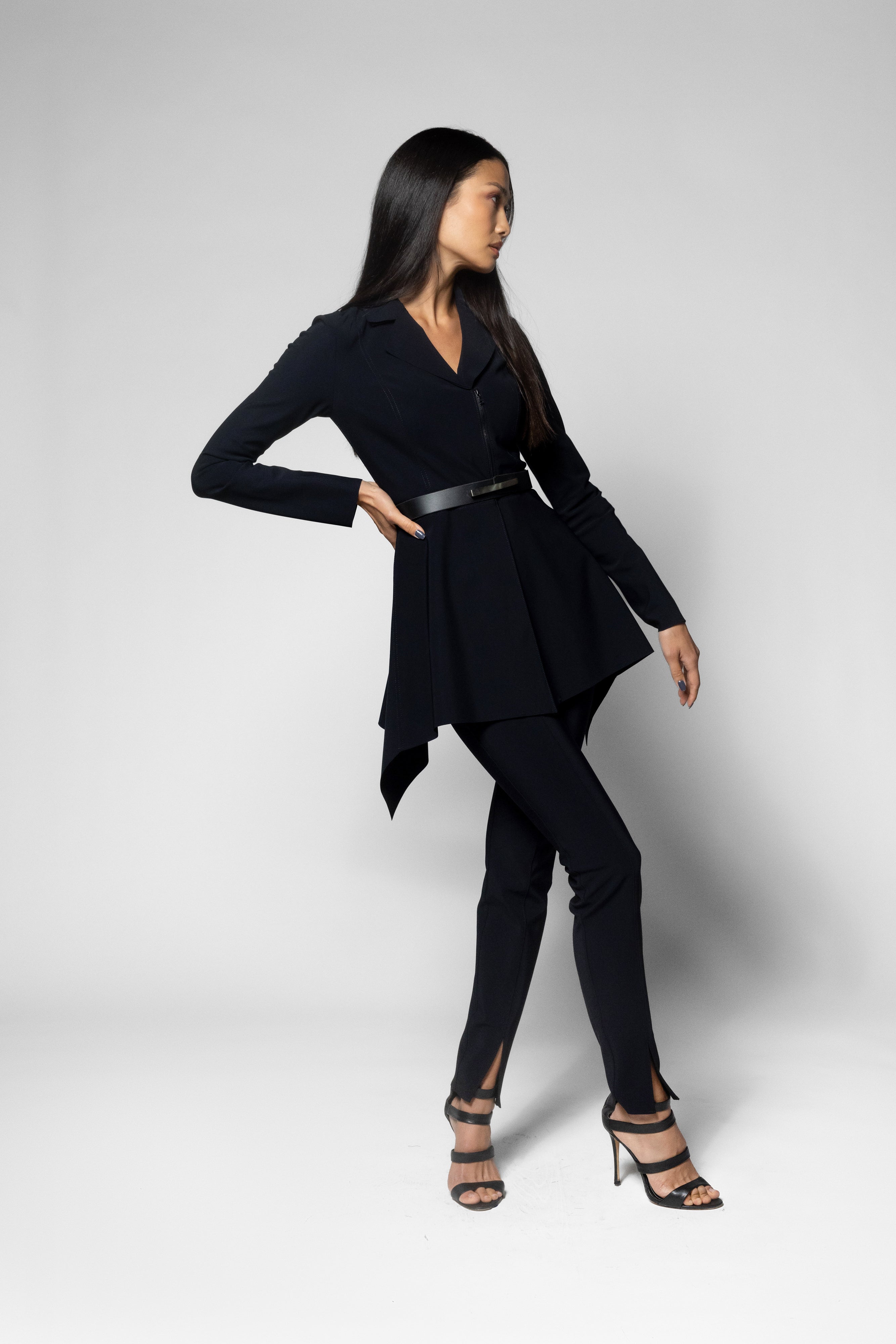 A woman modeling a black long-sleeve wrap-style top with a peplum hem, paired with black pants and strappy high-heeled sandals, against a light gray studio background.