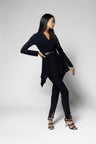 A woman modeling a black long-sleeve wrap-style top with a peplum hem, paired with black pants and strappy high-heeled sandals, against a light gray studio background.