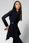 A woman modeling a black long-sleeve blazer-style top with a structured collar, belted waist, and layered asymmetrical hem, paired with black pants against a light gray studio background.