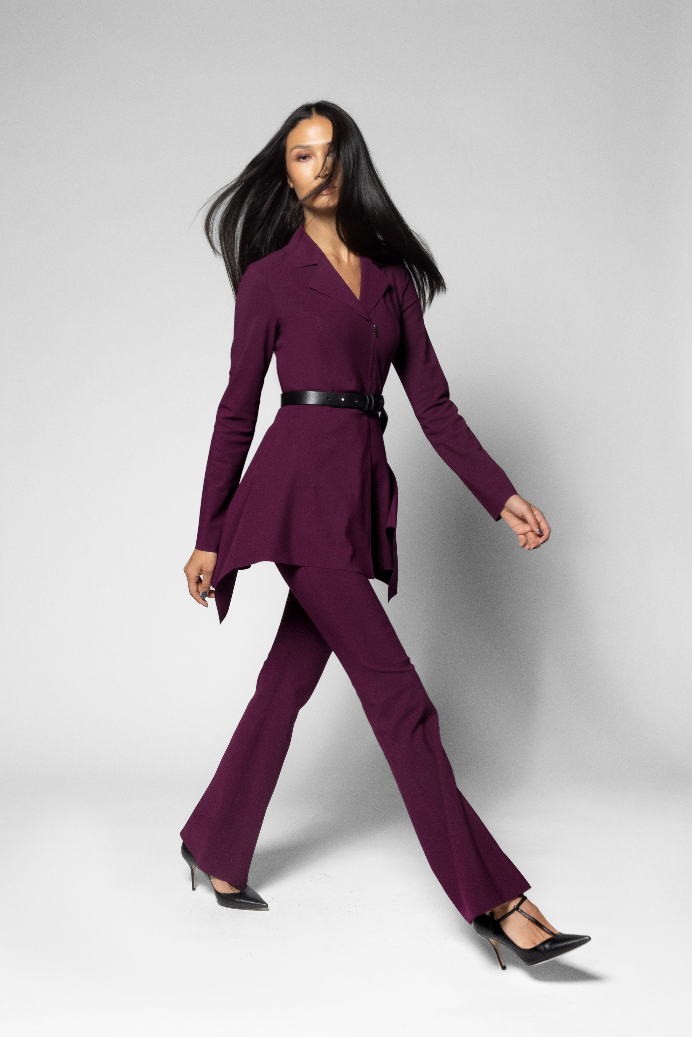 A woman modeling a deep purple two-piece suit with a wrap-style blazer and matching flared pants, cinched at the waist with a black belt, against a light gray studio background.