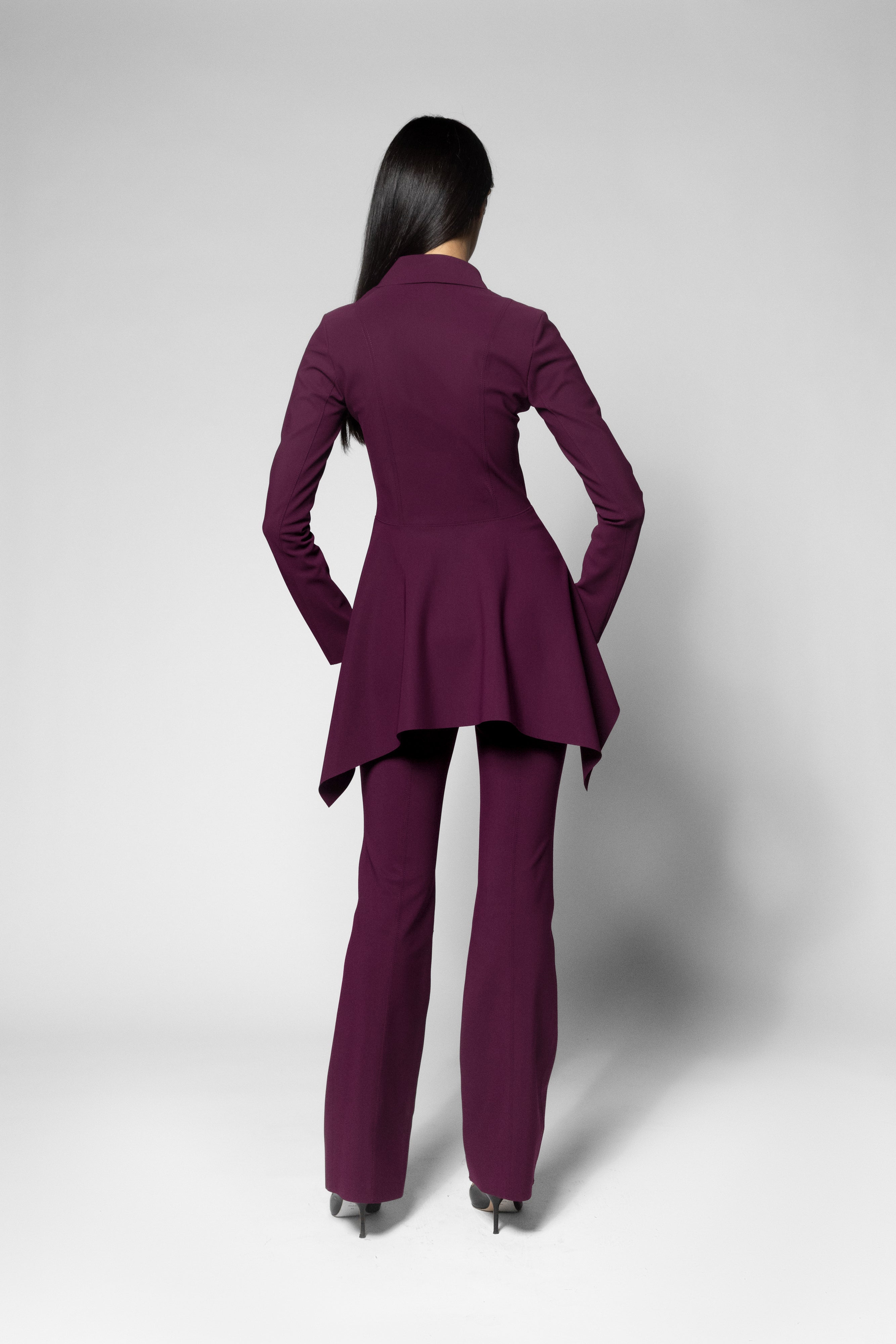 A model viewed from the back wearing a matching deep plum purple pantsuit, featuring a long-sleeve peplum-style blazer and matching trousers, set against a plain light gray studio background.