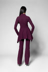 A model viewed from the back wearing a matching deep plum purple pantsuit, featuring a long-sleeve peplum-style blazer and matching trousers, set against a plain light gray studio background.