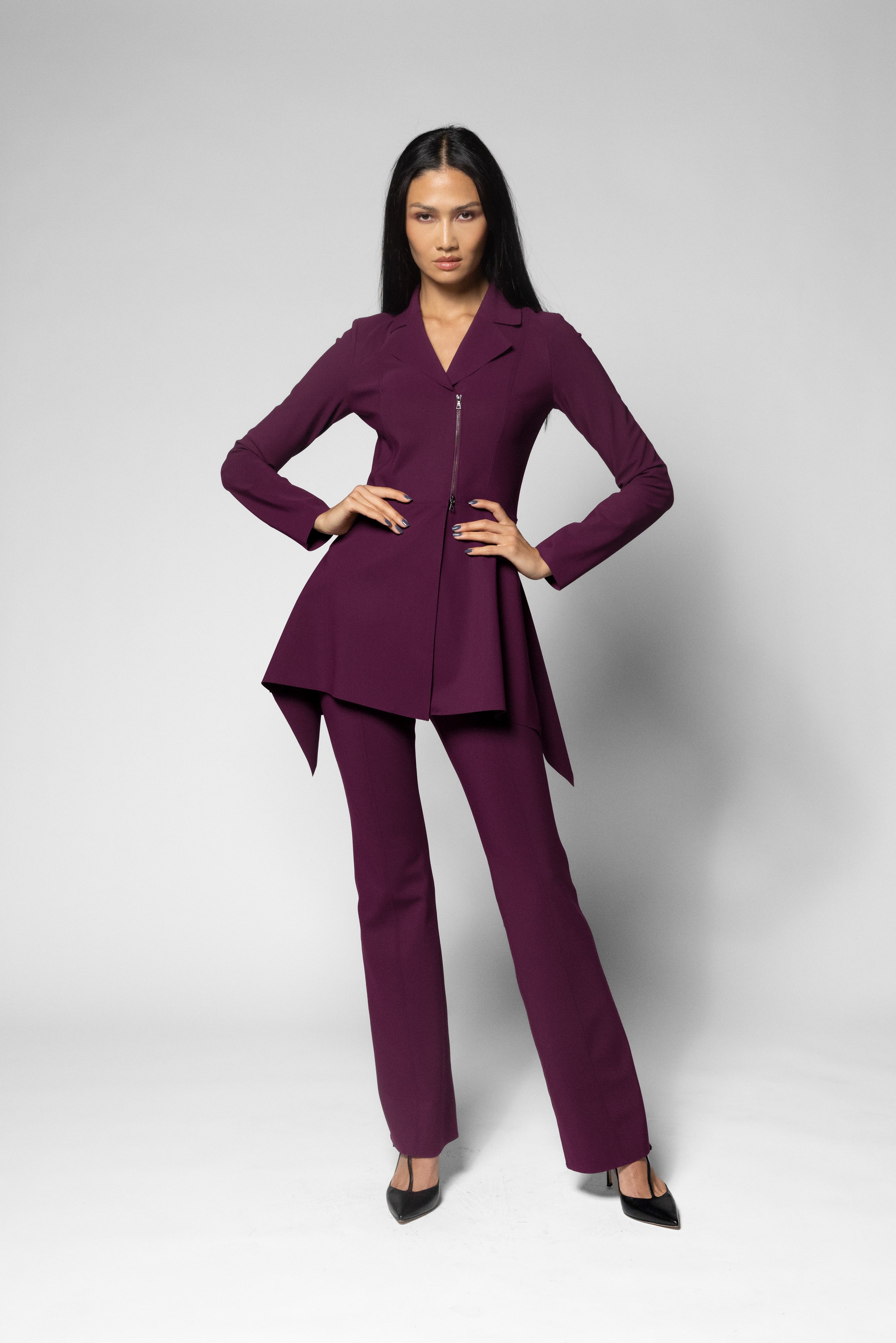 A woman modeling a matching deep purple two-piece suit consisting of a long-sleeve peplum-style jacket with a front zipper and matching straight-leg trousers, posed against a light gray studio background.