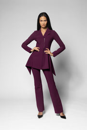 A woman modeling a matching deep purple two-piece suit consisting of a long-sleeve peplum-style jacket with a front zipper and matching straight-leg trousers, posed against a light gray studio background.