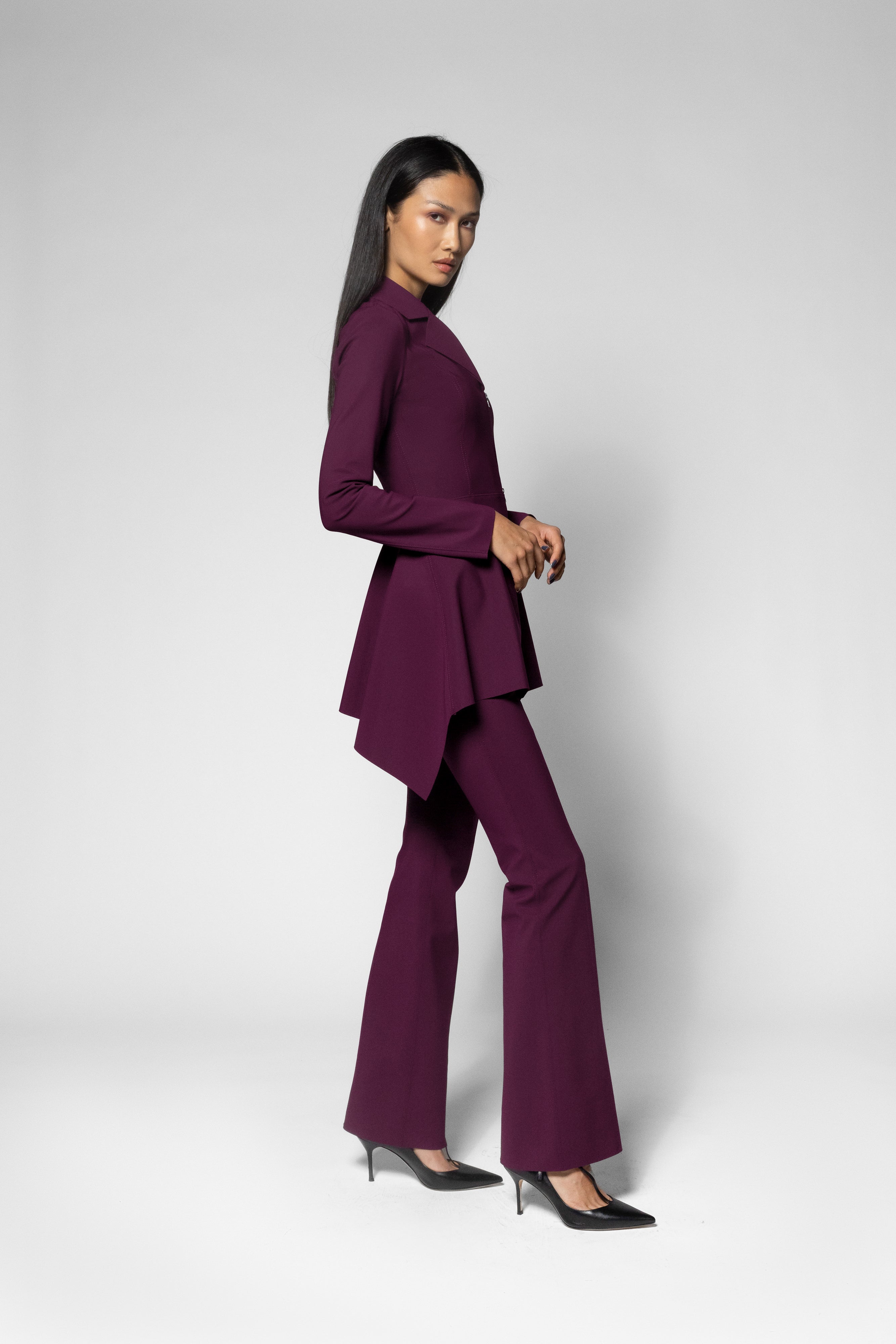 A model wearing a deep purple two-piece suit consisting of a long-sleeve, asymmetrical draped top and matching wide-leg pants, paired with black pointed-toe heels, against a light gray studio background.