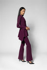 A model wearing a deep purple two-piece suit consisting of a long-sleeve, asymmetrical draped top and matching wide-leg pants, paired with black pointed-toe heels, against a light gray studio background.