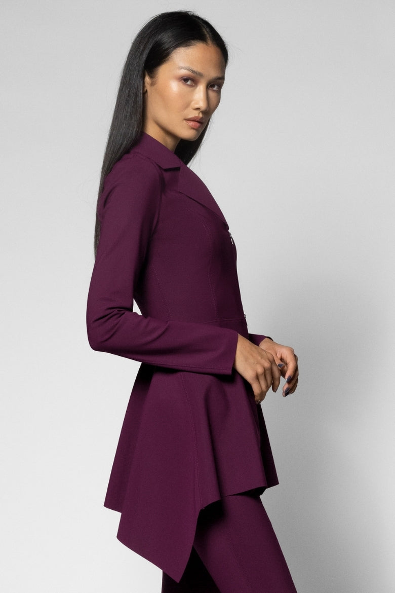A woman modeling a deep plum purple long-sleeve blazer with a high-low asymmetrical hem, shown from a side-back angle against a light gray studio background.