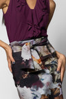 A close-up of a model wearing a sleeveless deep purple ruffled top and a high-waisted skirt with matching belt in a dark abstract floral print in black, white, and gray.