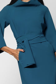 A close-up of a woman wearing a teal blue dress with a high mock neck, three-quarter sleeves, and a matching fabric tie belt at the waist.