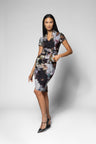 A woman modeling a short-sleeve collared knee-length dress and matching belt with a dark floral and abstract print in black, gray, white, and muted blue tones, paired with black pointed heels, against a light gray studio background.