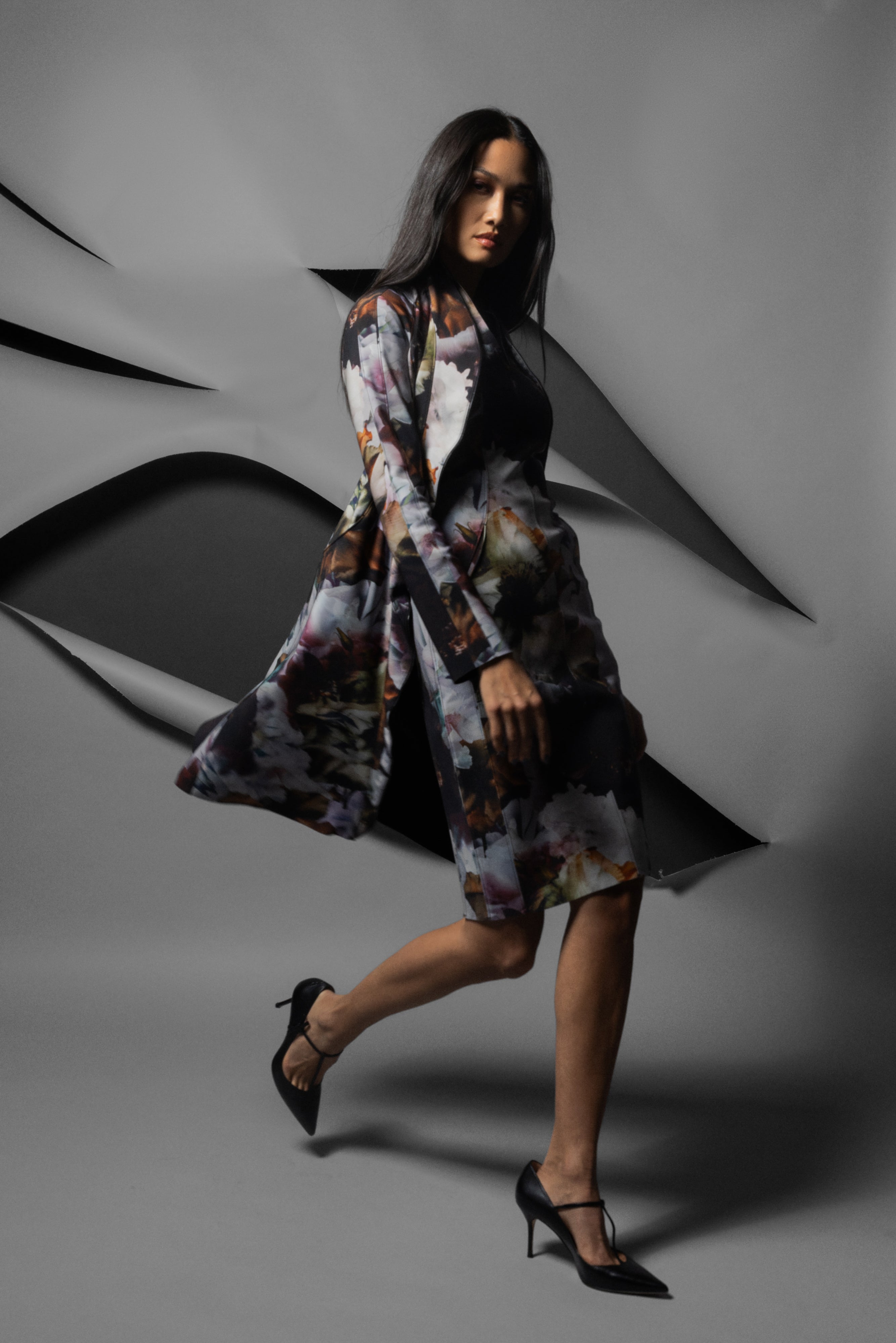 A woman models a long-sleeved, knee-length floral print wrap or coat over a black dress, paired with black pointed-toe heels, set against a stylized gray background with abstract layered paper-like shapes.