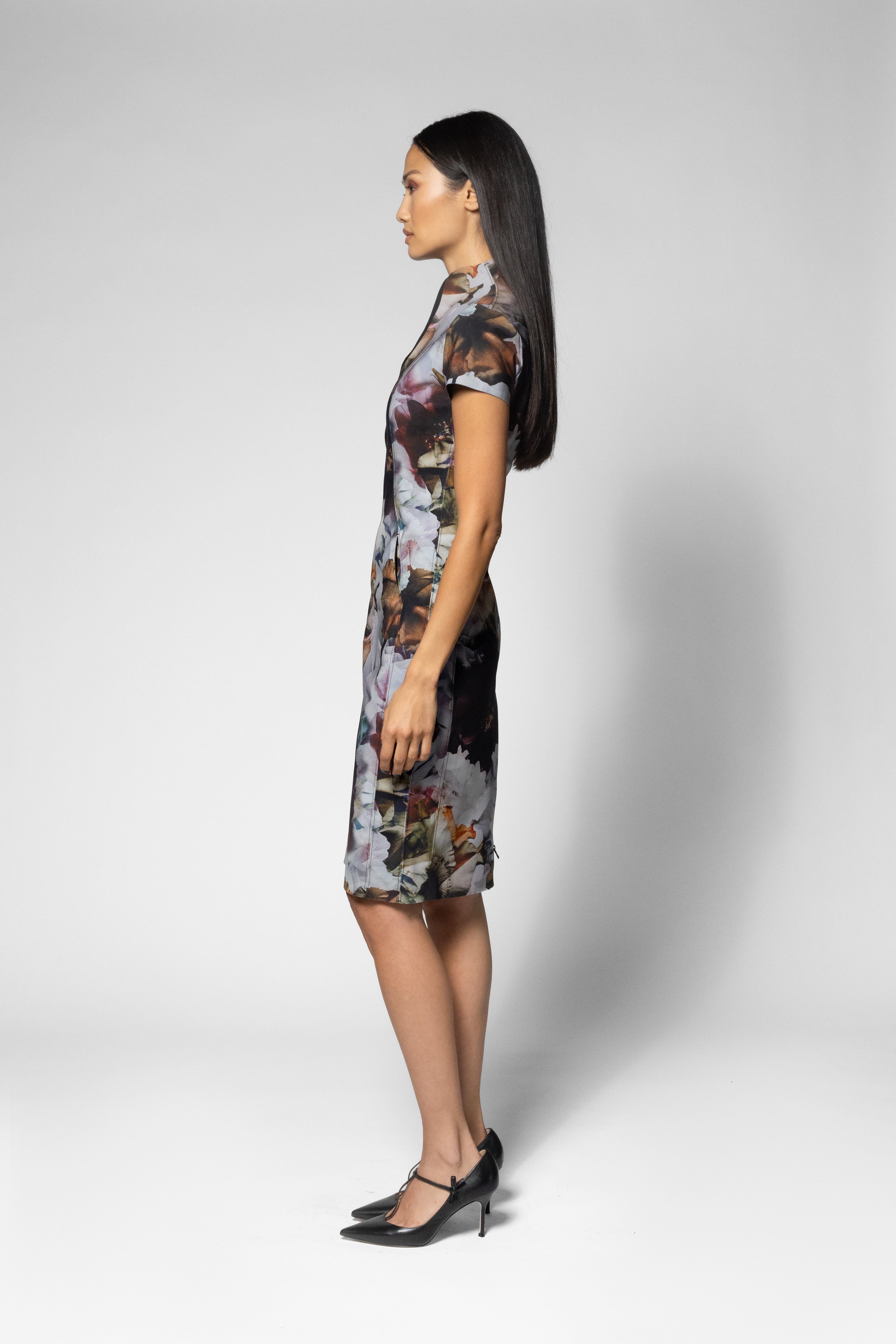 A woman in profile wearing a short-sleeved, knee-length dress with a dark base and a painterly floral pattern in muted gray, brown, and white tones, paired with black pointed-toe heels, against a light gray studio background.