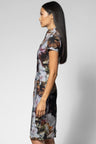 A woman shown in side profile wearing a short-sleeve cheongsam-style dress with a muted floral print in gray, brown, beige, and white tones against a light gray studio background.