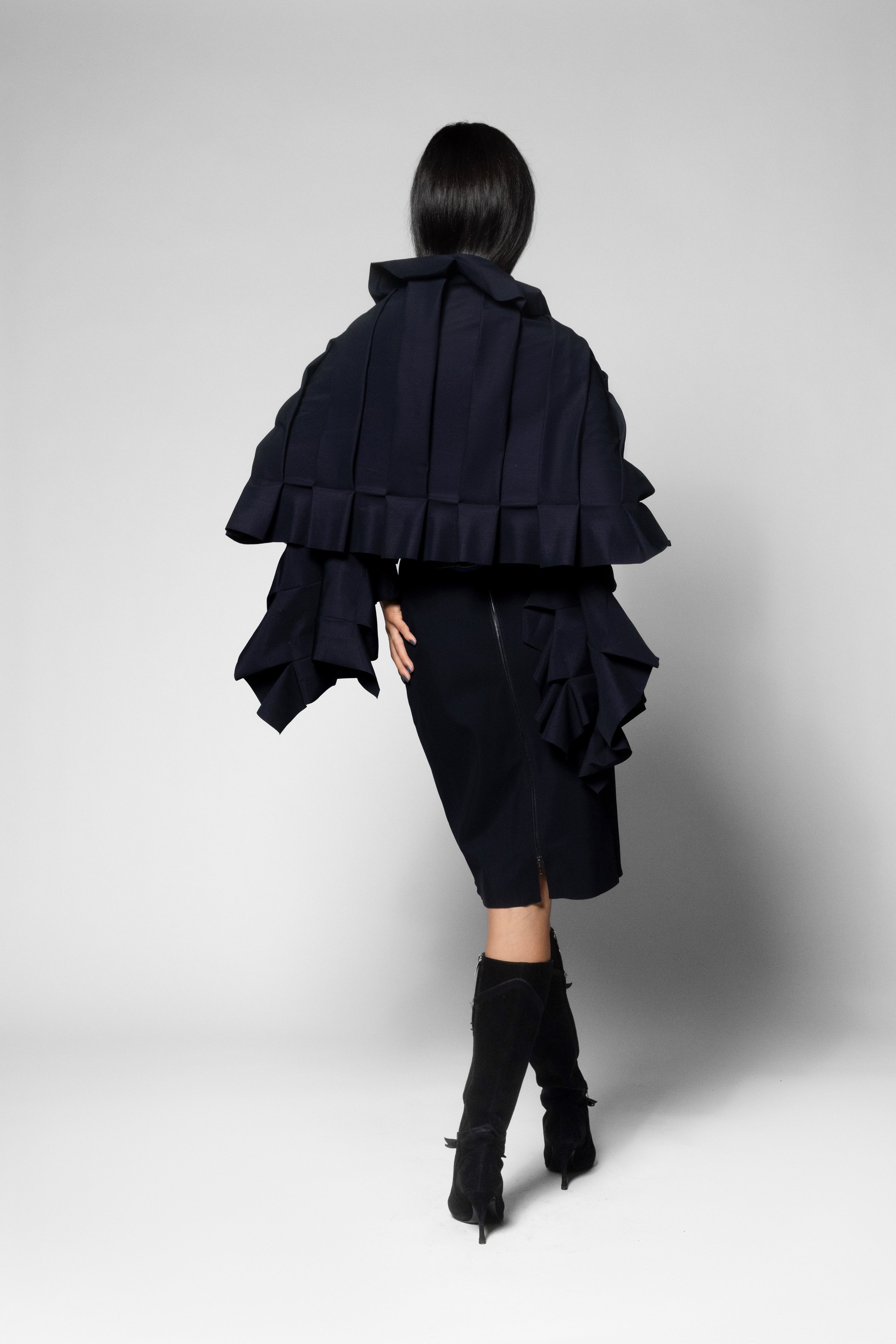 A model shown from the back wearing a dark navy ruffled cape-style scarf with dramatic layered pleats, a matching black dress, and black knee-high boots against a light gray studio background.
