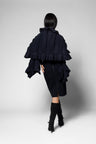 A model shown from the back wearing a dark navy ruffled cape-style scarf with dramatic layered pleats, a matching black dress, and black knee-high boots against a light gray studio background.