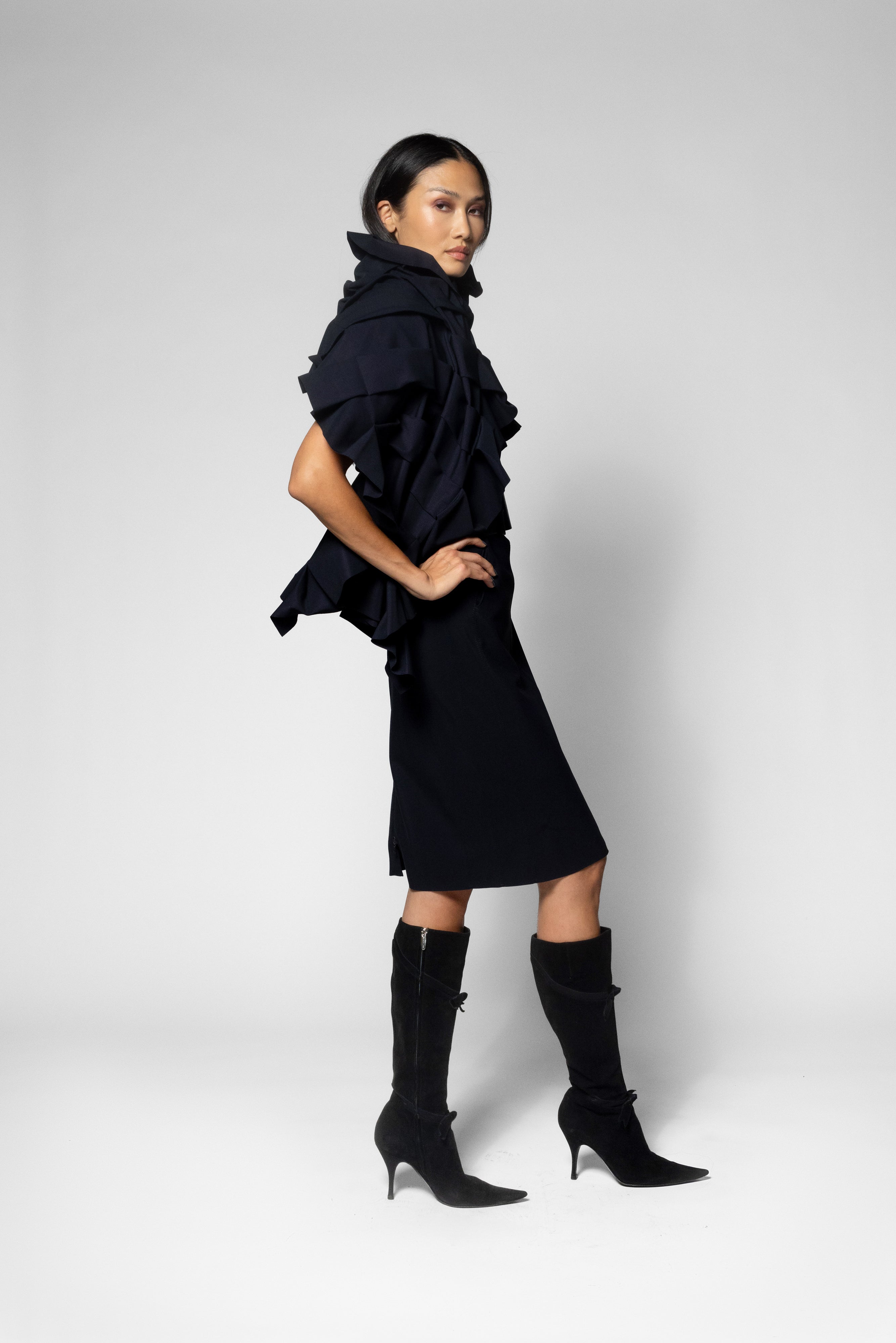 A model wearing a black ruffled scarf over short-sleeve dress, paired with black knee-high heeled boots, photographed against a light gray studio background.