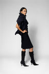 A model wearing a black ruffled scarf over short-sleeve dress, paired with black knee-high heeled boots, photographed against a light gray studio background.