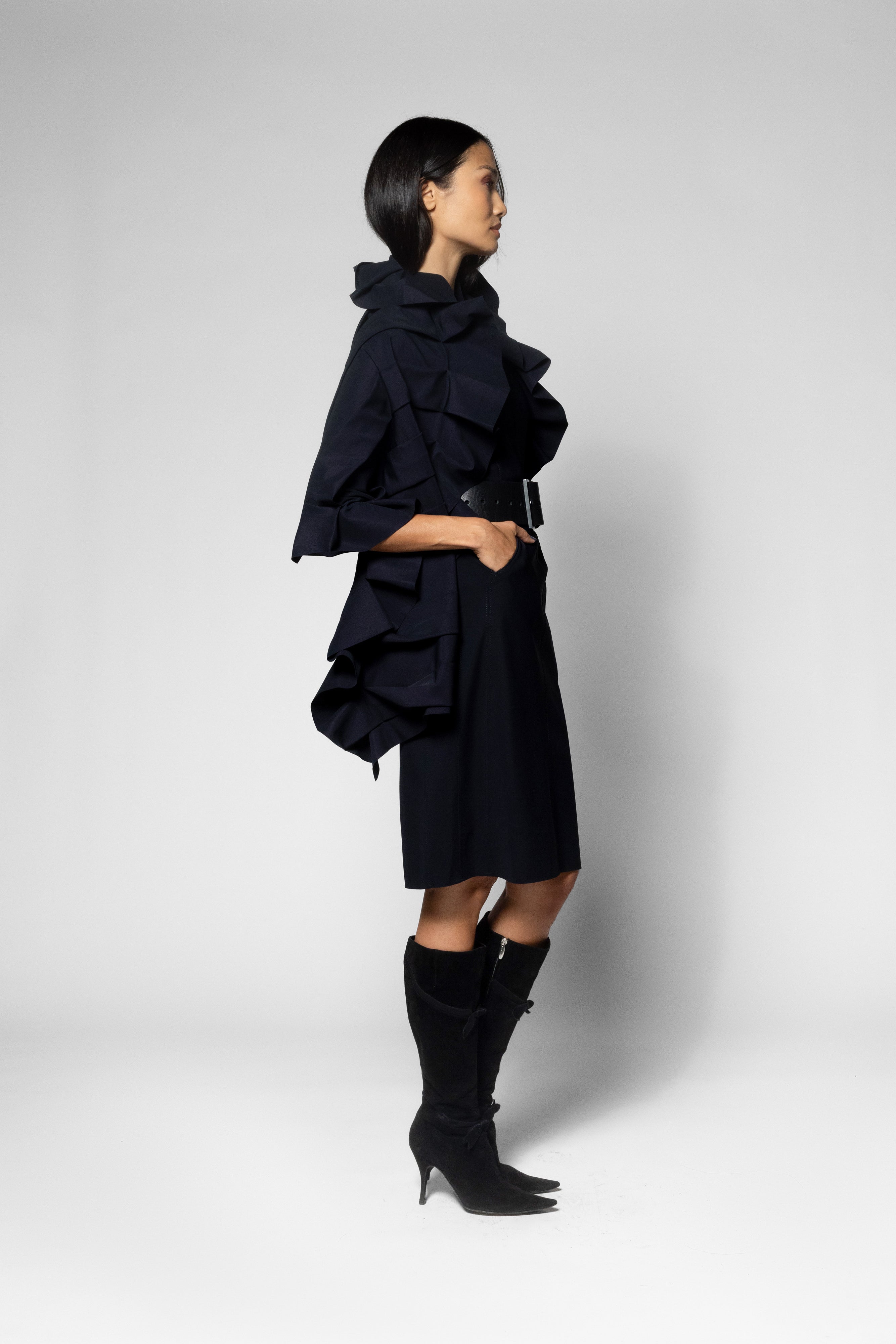 A model in profile wearing a black ruffled scarf with a high collar, a matching midi dress, and tall black heeled boots, photographed against a light gray studio background.