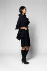 A model in profile wearing a black ruffled scarf with a high collar, a matching midi dress, and tall black heeled boots, photographed against a light gray studio background.