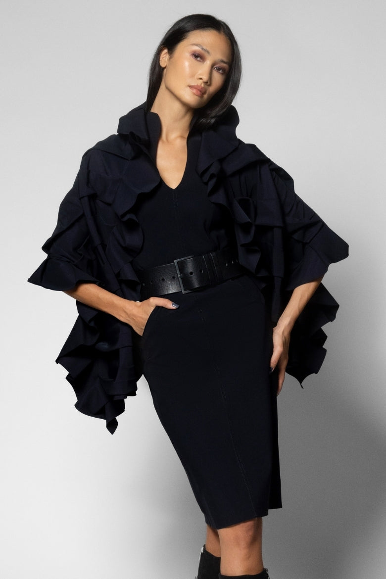 A model wearing a black ruffled cape-style wrap over a black V-neck dress with a wide black belt, photographed against a light gray studio background.