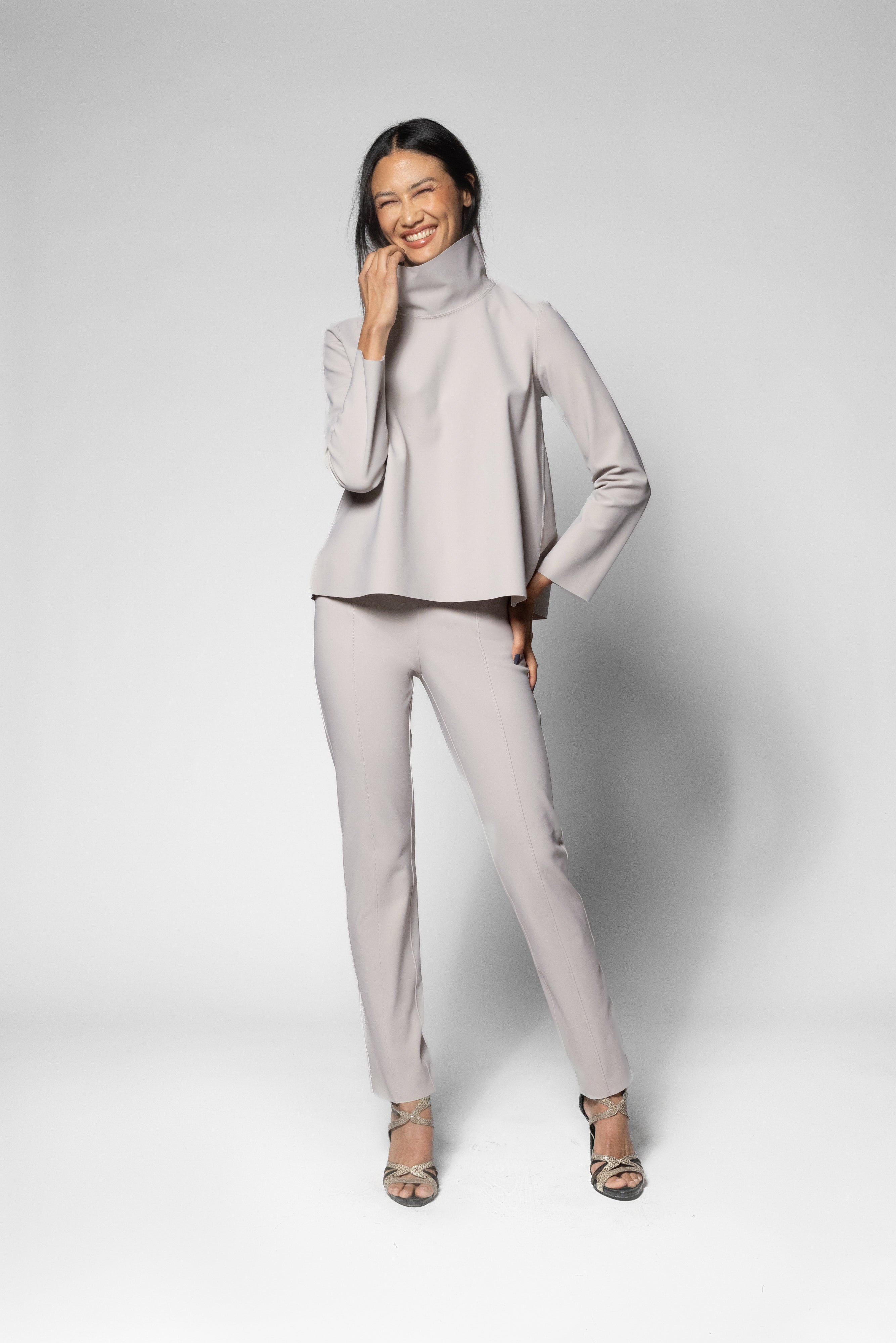 A woman modeling a light gray two-piece outfit consisting of a long-sleeve mock-neck top and matching slim-fit pants, paired with high heels, against a light gray studio background.