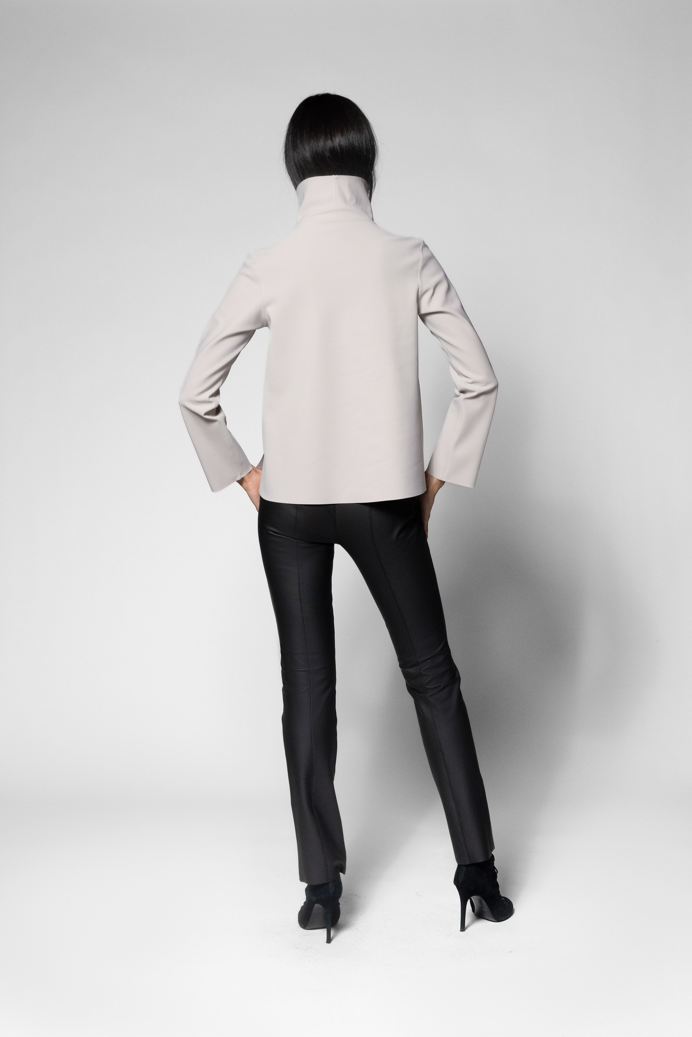 A model shown from the back wearing a light beige mock-neck long-sleeve top and black pants, standing against a light gray studio background.