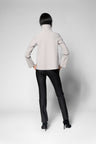 A model shown from the back wearing a light beige mock-neck long-sleeve top and black pants, standing against a light gray studio background.