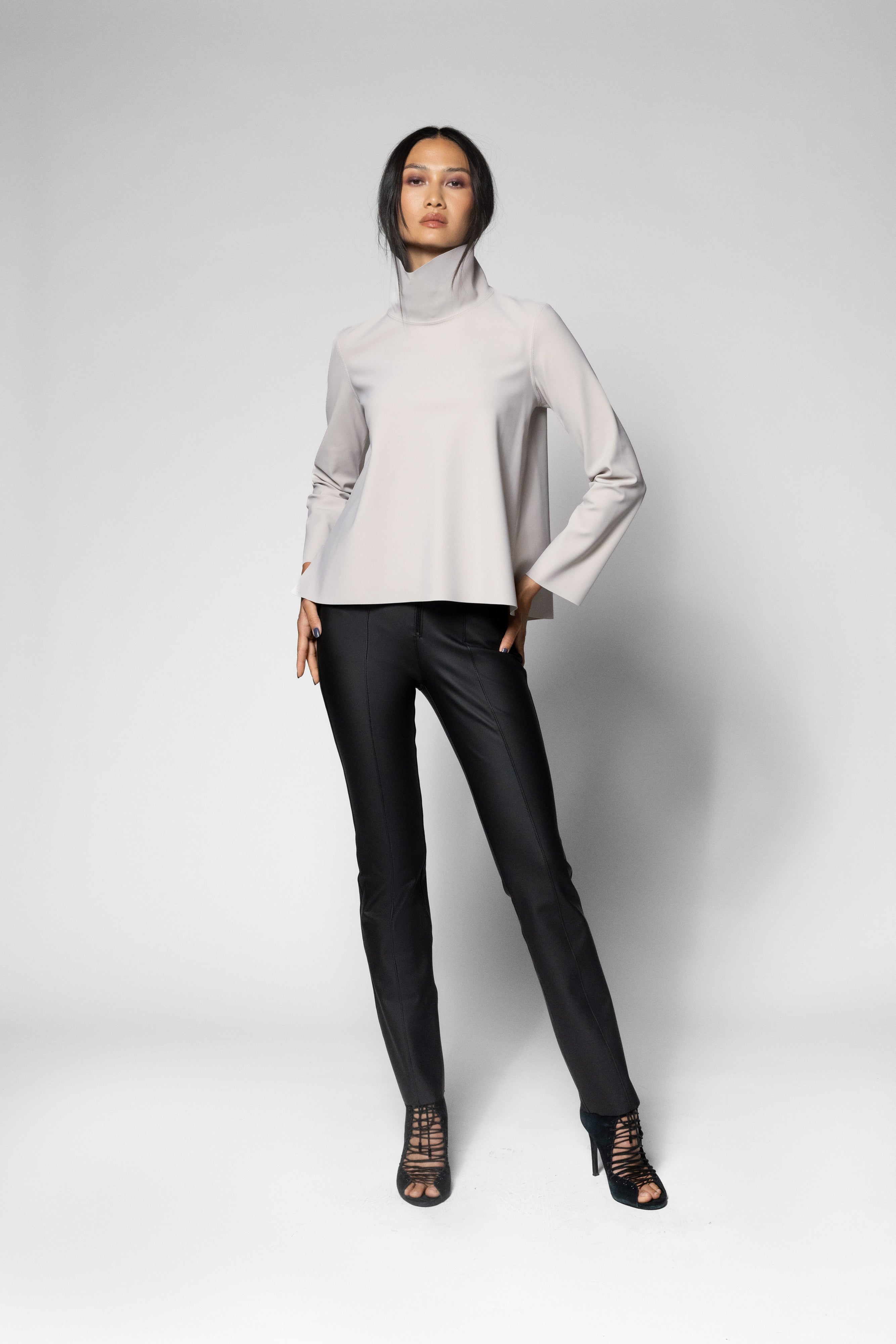 A model stands against a light grey studio background wearing a light beige long-sleeve mock-neck top, black pants, and black lace-up high-heeled sandals.