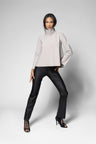 A model posing in a studio setting, wearing a light beige high-neck long-sleeve top, black trousers, and black lace-up high heels against a light gray background.