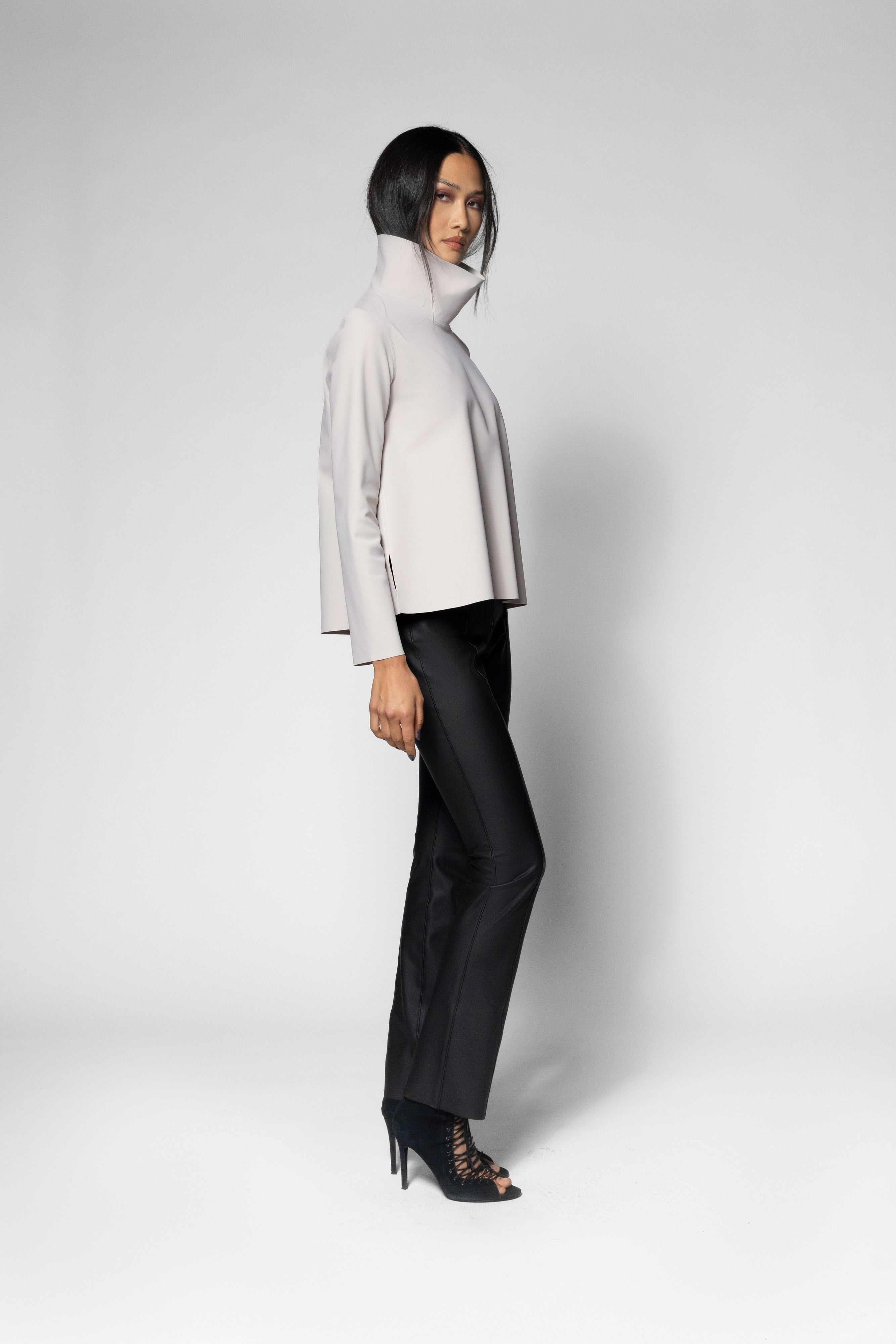 A model wearing a light beige high-neck long-sleeve top, black trousers, and black lace-up high heels, standing in profile against a light gray studio background.
