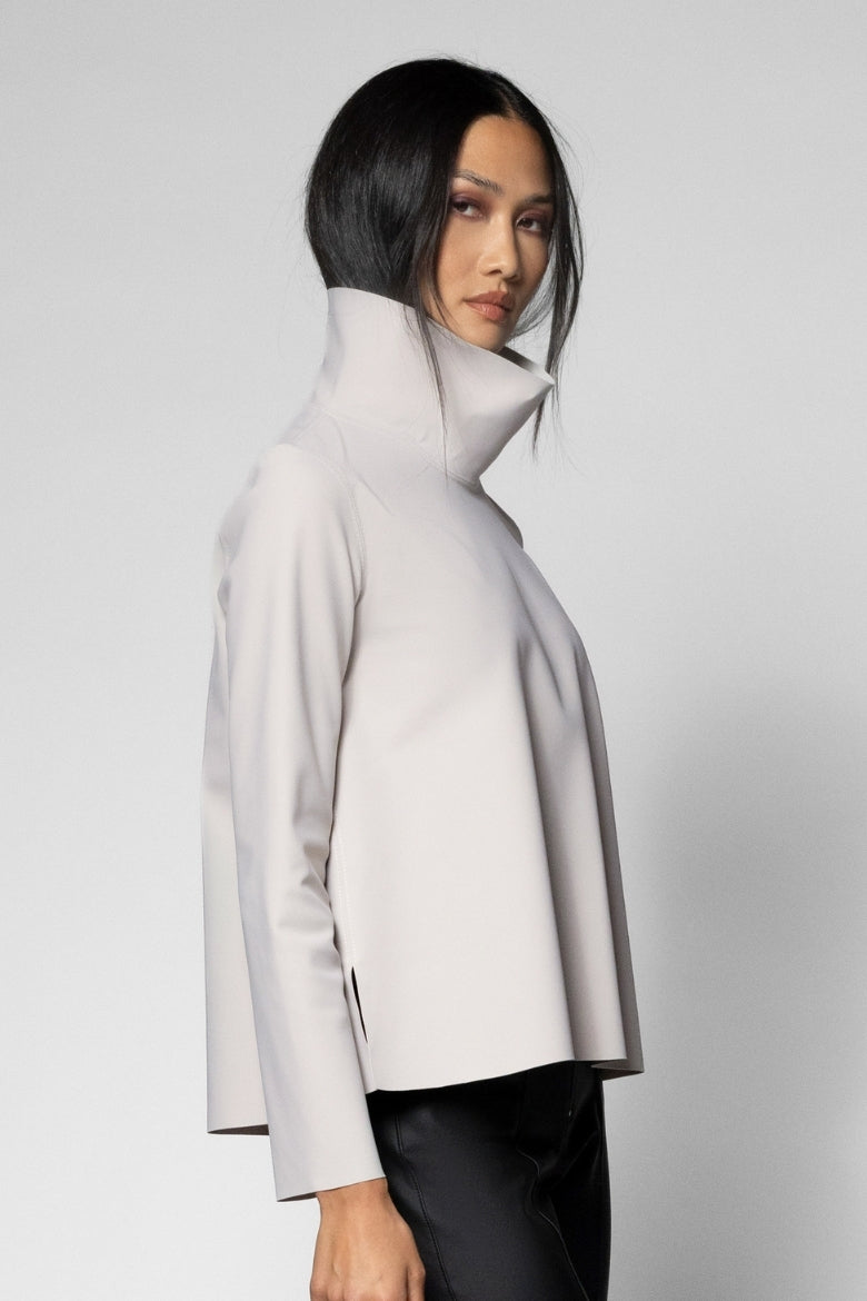 A model wearing a light gray high-neck, long-sleeve structured jacket with black pants, photographed against a neutral light gray studio background.