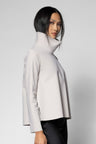 A model wearing a light gray high-neck, long-sleeve structured jacket with black pants, photographed against a neutral light gray studio background.