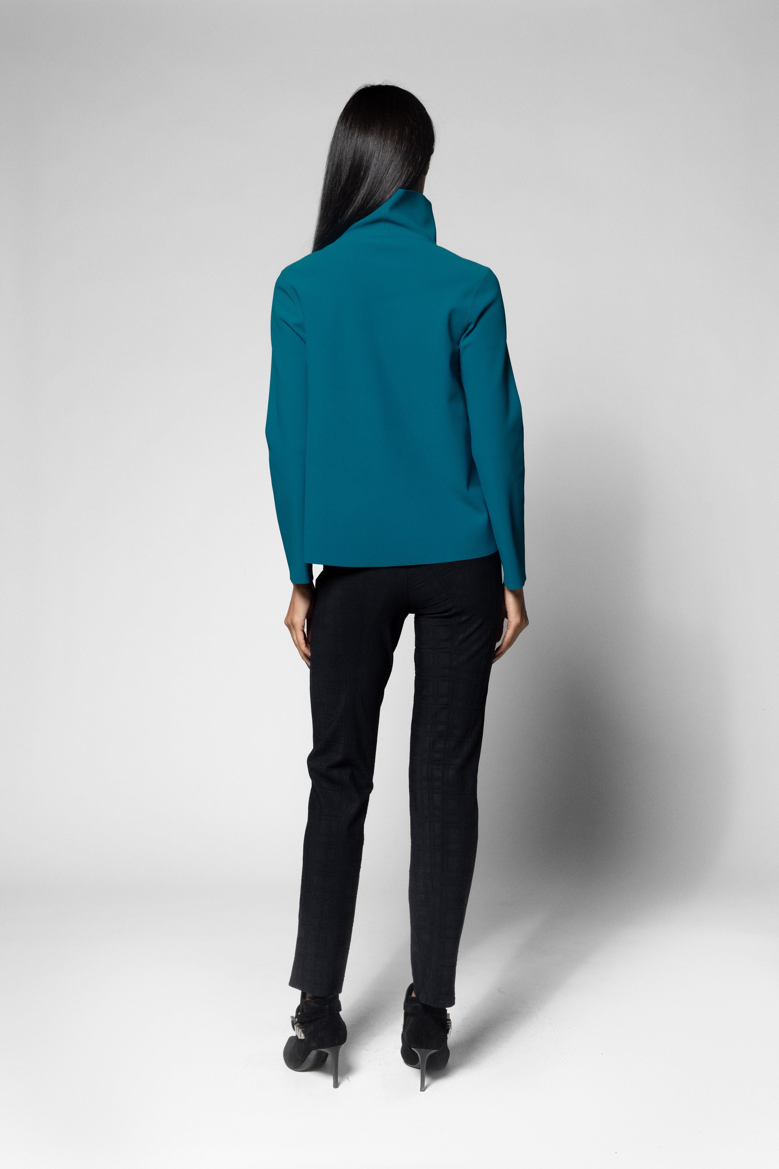 A woman is shown from the back wearing a teal long-sleeve turtleneck, black pants, and black high-heeled shoes against a light gray studio background.