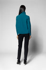A woman is shown from the back wearing a teal long-sleeve turtleneck, black pants, and black high-heeled shoes against a light gray studio background.