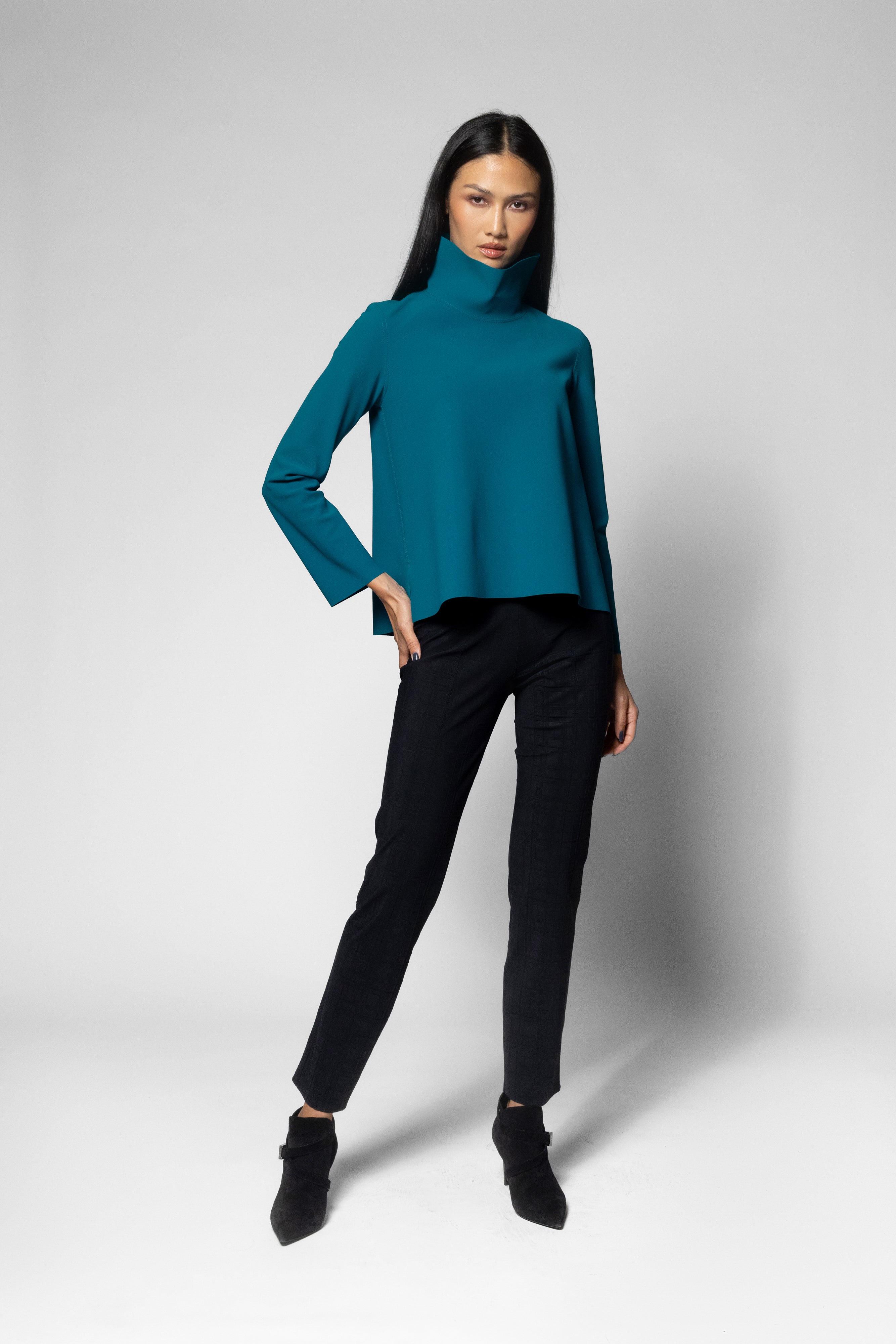 A woman modeling a teal long-sleeve mock neck top with black pants and black ankle boots, standing against a light gray studio background.