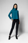 A woman modeling a teal long-sleeve mock neck top with black pants and black ankle boots, standing against a light gray studio background.