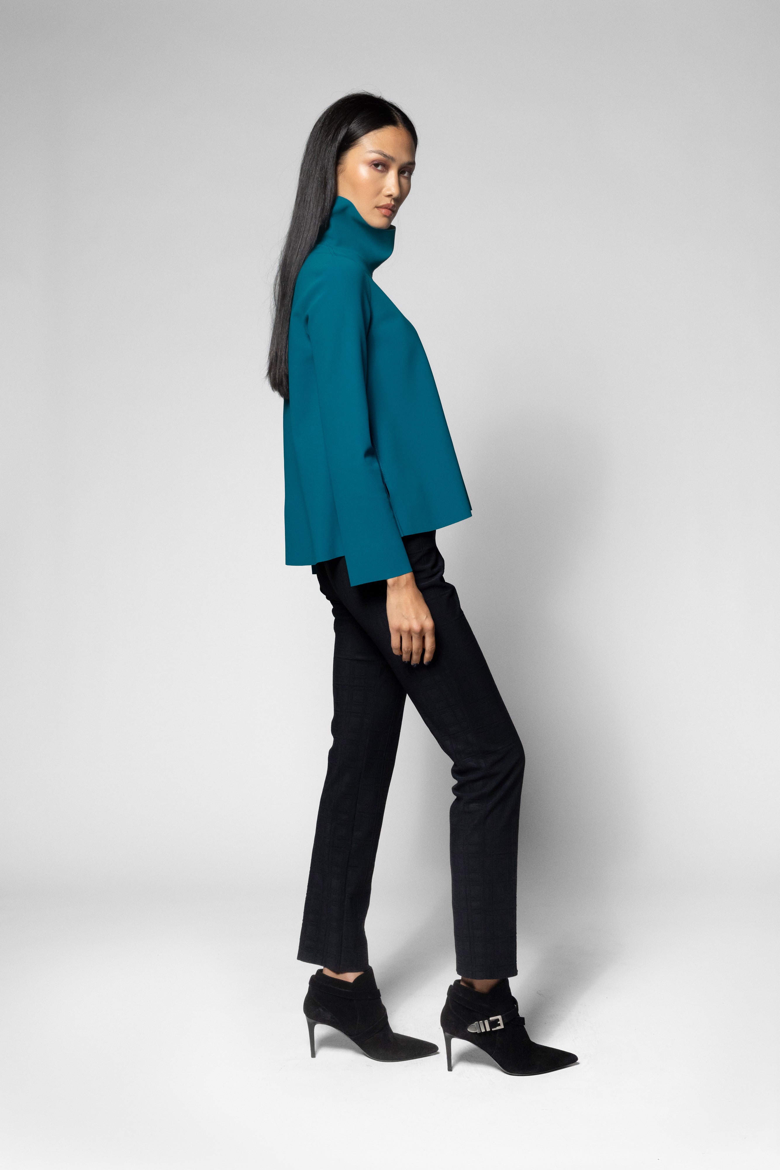 A woman modeling a teal turtleneck top, black trousers, and black ankle boots with silver buckle details, posed against a light gray studio background.