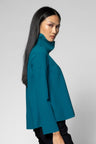 A woman with long, straight black hair is shown in profile wearing a teal high-neck cape-style top and black pants against a plain light gray studio background.