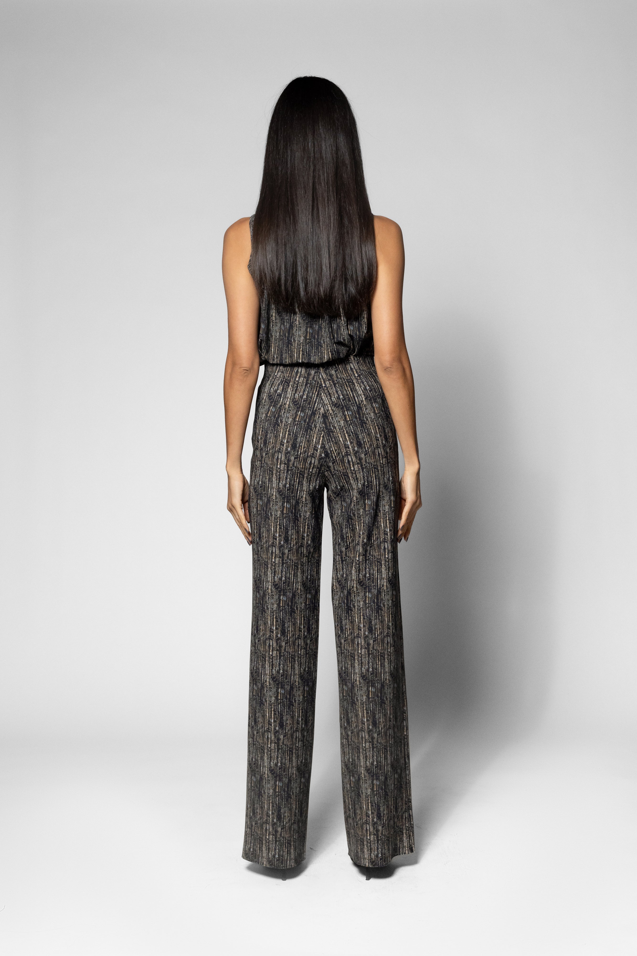 A woman viewed from the back wearing a sleeveless, high-waisted jumpsuit with wide legs in a textured black and gray pattern, set against a plain light gray studio background.