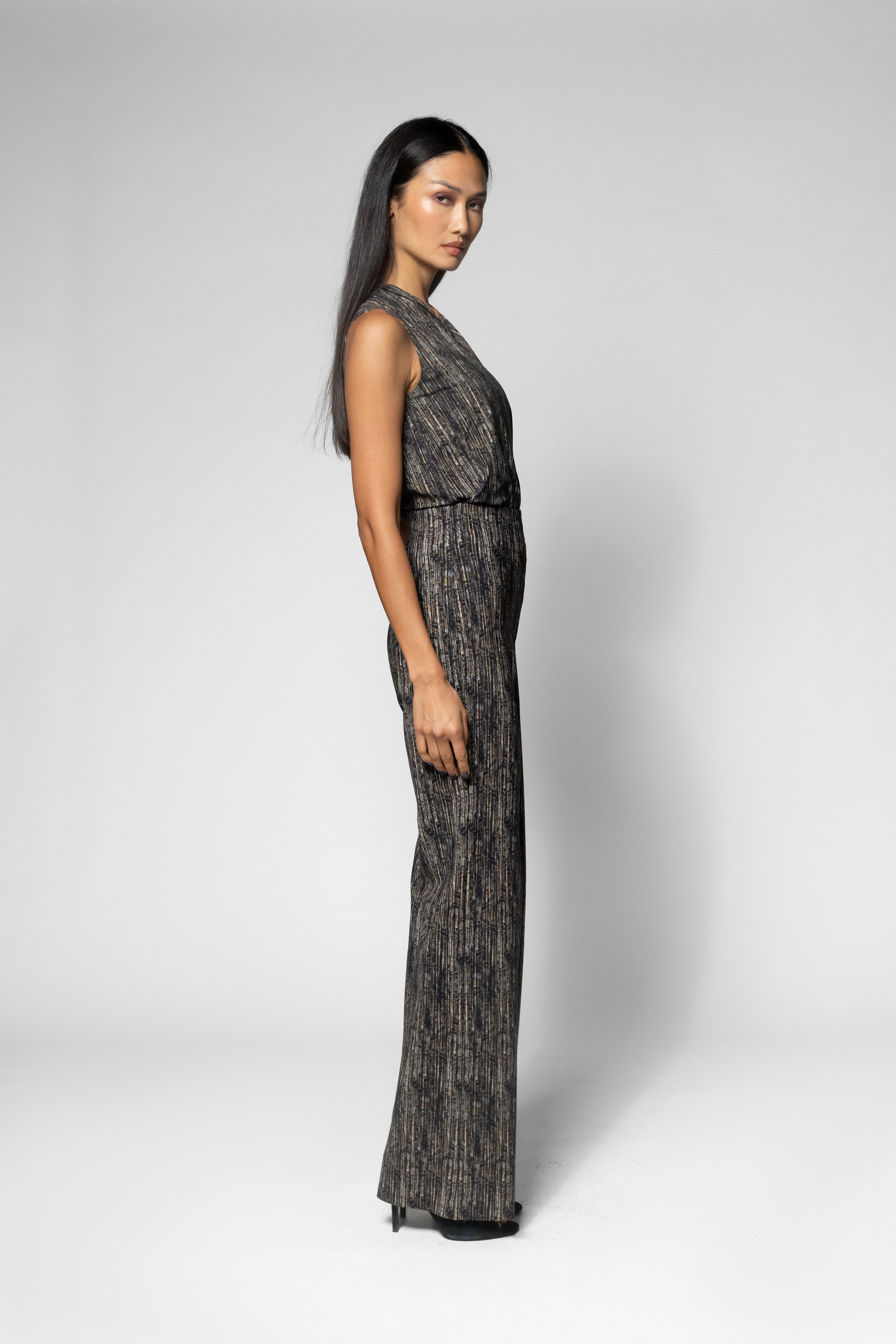 A woman modeling a sleeveless, floor-length dark gray and black textured jumpsuit against a light gray studio background.