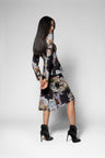 A woman modeling a long-sleeve midi dress with a dark abstract floral print, paired with black ankle boots, against a light gray studio background.