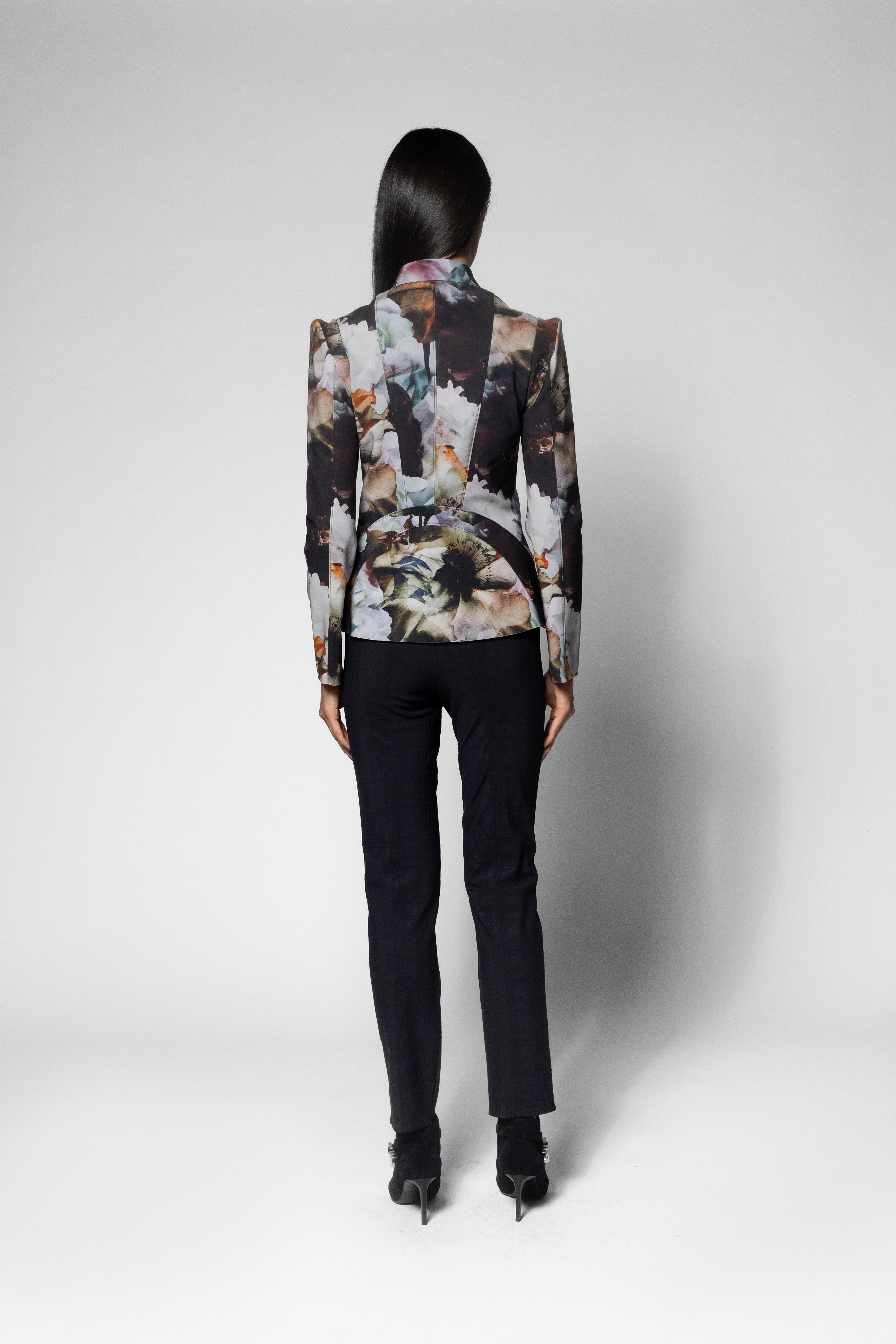 A model shown from the back wearing a structured blazer with a complex abstract floral print in black, white, beige, and brown, paired with black trousers and heels, against a light gray studio background.