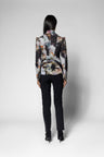 A model shown from the back wearing a structured blazer with a complex abstract floral print in black, white, beige, and brown, paired with black trousers and heels, against a light gray studio background.