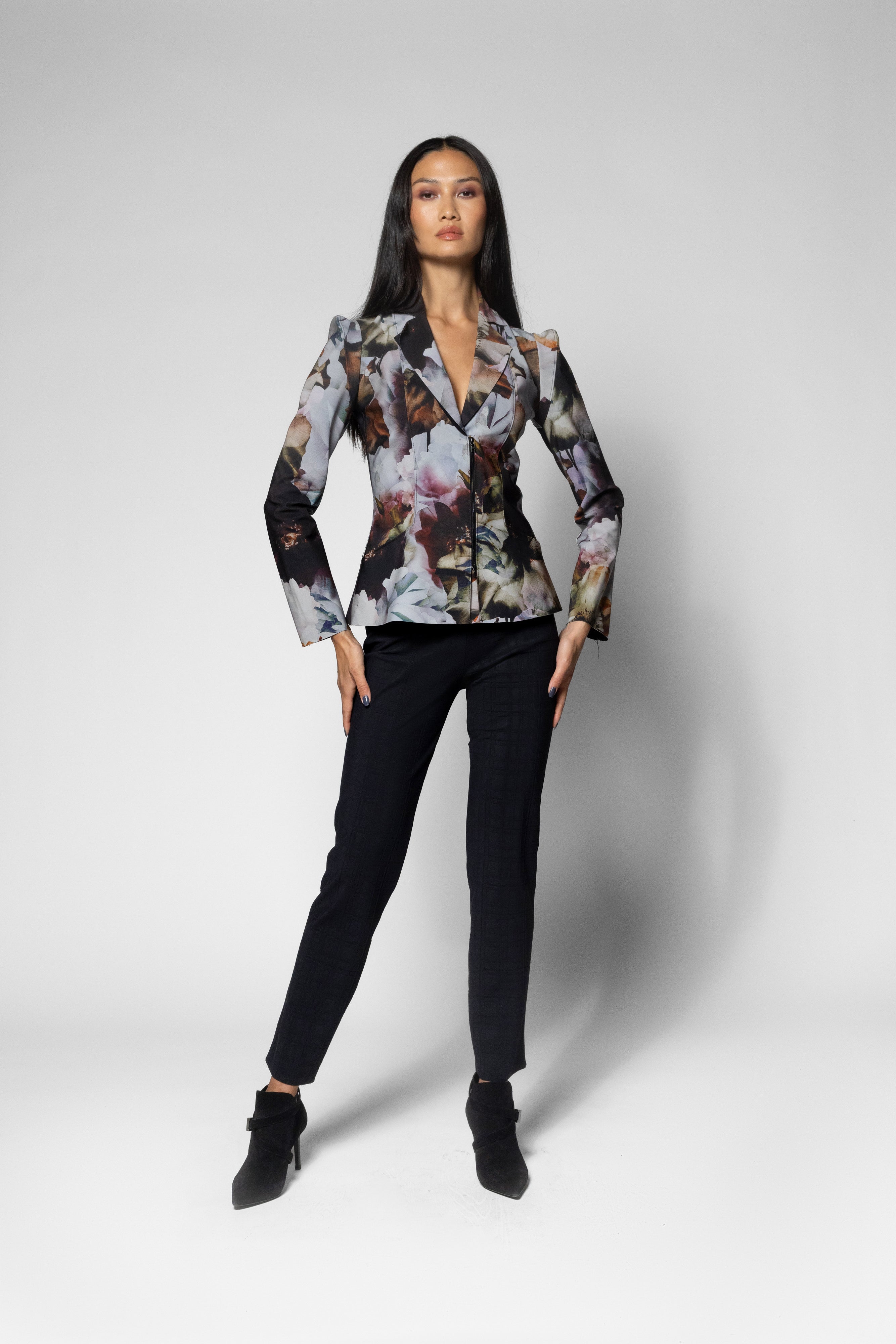 A woman modeling a long-sleeve jacket with a complex multicolor floral and abstract print, paired with black trousers and black ankle boots, against a light gray studio background.
