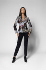 A woman modeling a long-sleeve jacket with a complex multicolor floral and abstract print, paired with black trousers and black ankle boots, against a light gray studio background.